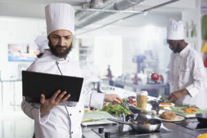 In a restaurant kitchen, male chef in a uniform holds a laptop researching recipes while doing food prep with vegetables and other ingredients. Cooking gourmet dishes on the stove.